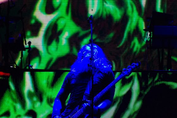 Musician playing bass guitar on stage with vibrant green and blue background lighting.