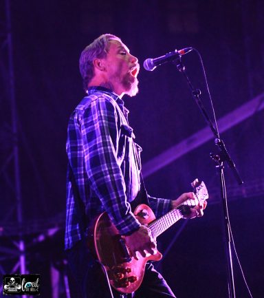 Musician performing on stage with a guitar, wearing a plaid shirt and singing into a microphone.