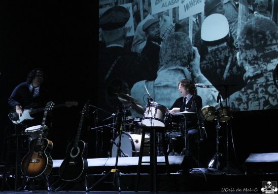 Musicians performing on stage with guitars and drums, featuring a vintage background image.