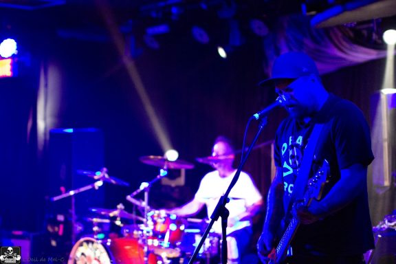 Musicians performing on stage with drums and guitar under blue stage lights.