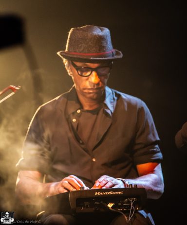 Musician in a black shirt and hat playing an instrument on stage with a smoky background.