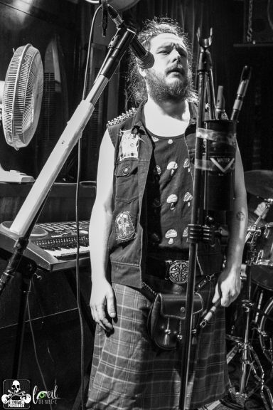 A musician in a kilt stands near microphones, performing with passion.