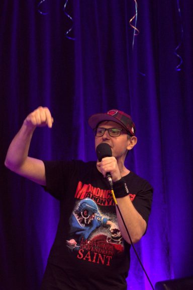 Person wearing a graphic t-shirt and cap, speaking into a microphone with a purple backdrop.