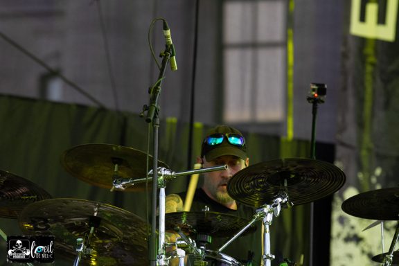 A drummer wearing sunglasses plays on stage, surrounded by drums and cymbals.
