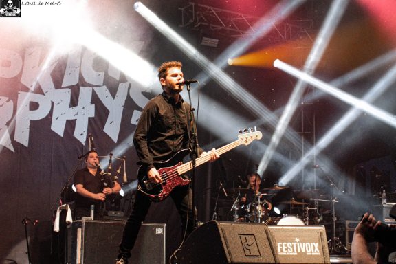 Musician performing on stage with a bass guitar, backed by a band and vibrant stage lights.