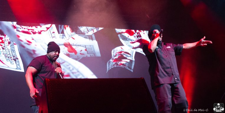 Two performers on stage, wearing black shirts and hats, with a colorful backdrop.