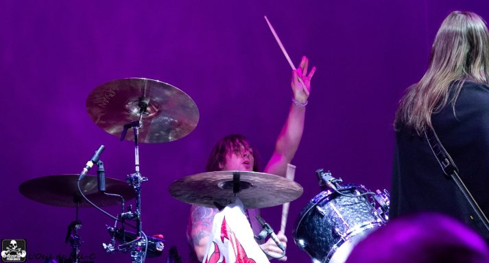 Drummer performing energetically on stage with a purple backdrop.
