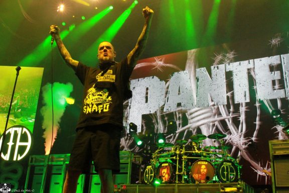 A performer with tattoos raises fists on stage in front of a large Pantera backdrop.