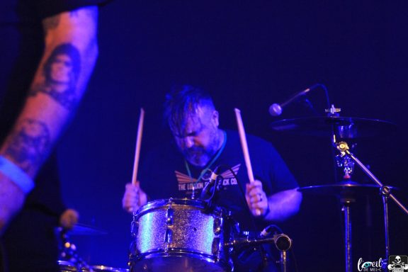 Drummer focused on playing, with a dark background and vibrant stage lighting.