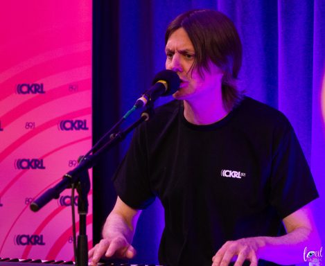 Musician performing at a keyboard with a microphone, under colorful stage lighting.