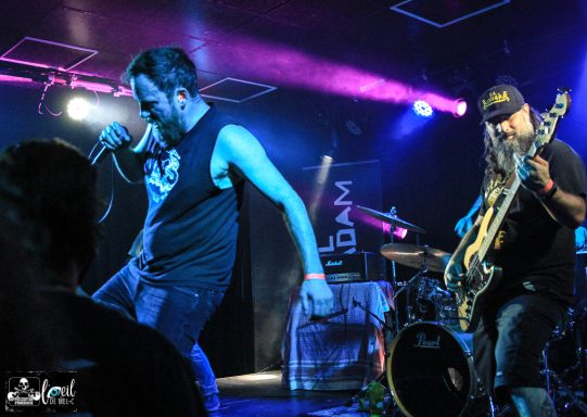 A vocalist performs energetically on stage while a bassist plays beside them.