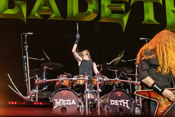 Drummer performing energetically on stage with vibrant lighting and band name backdrop.