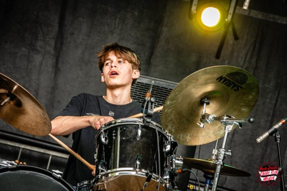 Young drummer playing energetically on stage, with focused expression.