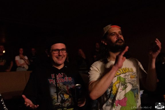 Two men in casual attire enjoying a live performance, smiling and clapping.