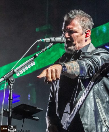 A musician with tattoos gestures emphatically while singing into a microphone on stage.