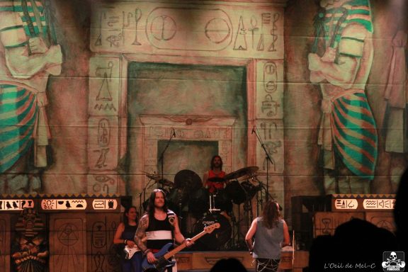 Band performing on stage with ancient-themed backdrop and large amplifiers.