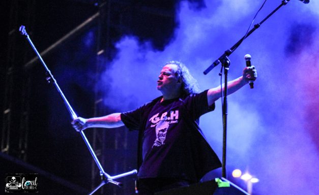 Musician performing on stage with a microphone, surrounded by purple and blue lighting.