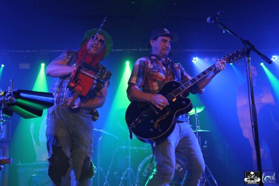 Two musicians perform on stage with instruments, dressed in colorful outfits under blue and green lights.
