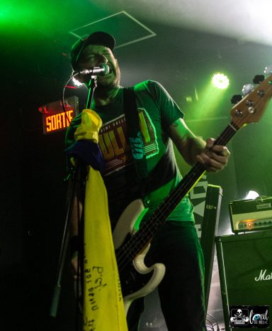 Musician playing bass on stage with green lighting and a colorful scarf.