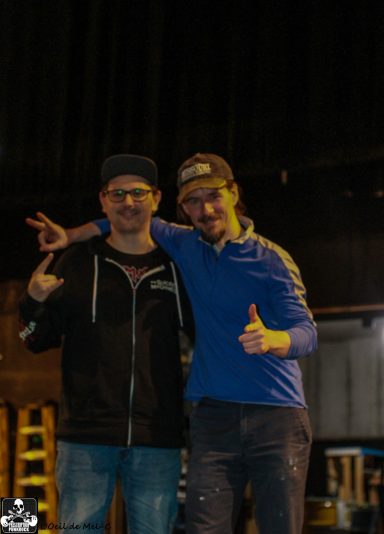 Two men stand together, smiling and posing with thumbs up in a stage setting.