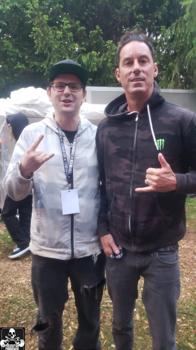 Two men standing outdoors, both making hand gestures and smiling at the camera.