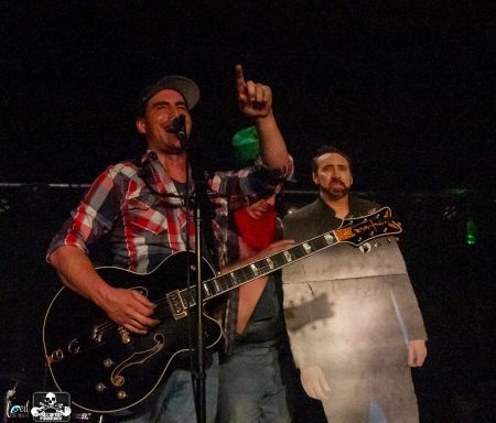 A musician on stage holding a guitar, pointing, with a life-sized cutout beside him.