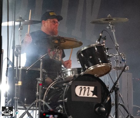 Drummer performing on stage with a black drum kit and a lit backdrop.