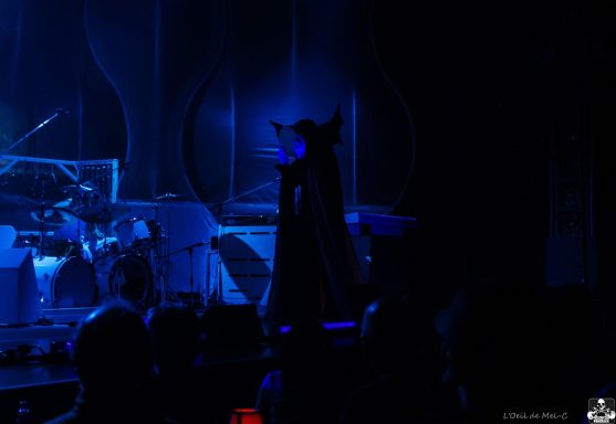 A figure in a dark cape stands against a blue-lit stage backdrop.