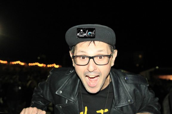 Person wearing glasses and a black cap, smiling enthusiastically against a dark background.
