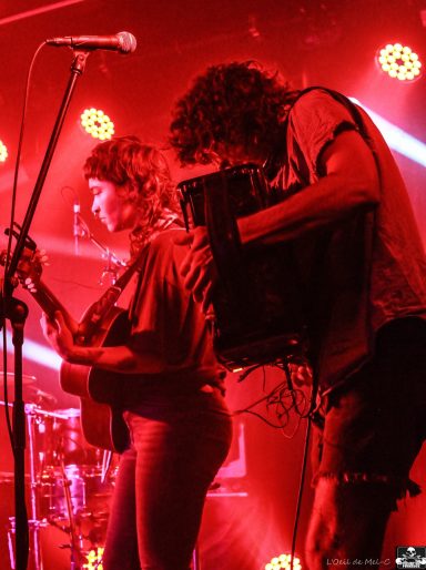 A musician plays accordion beside a singer performing on stage with red lighting.