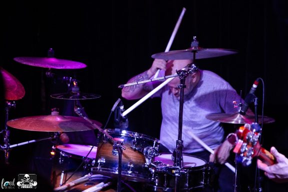 A drummer energetically playing with drumsticks against a dark background.