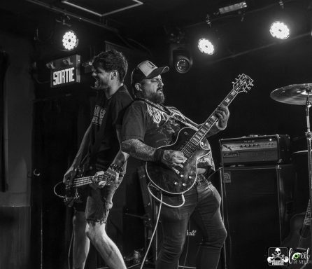 Two musicians perform on stage, one on guitar and the other on bass, in a lively setting.