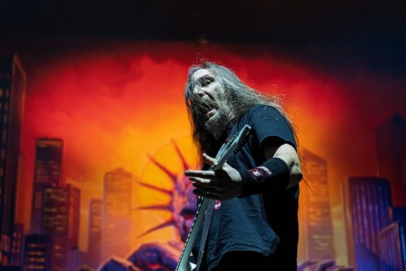 Man with long hair playing guitar on stage against a vibrant cityscape backdrop.